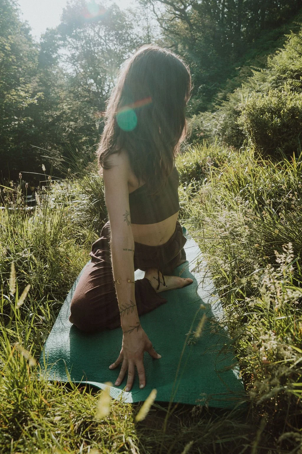 A woman practicing yoga outdoors on a mat in a grassy area, with trees and sunlight in the background.