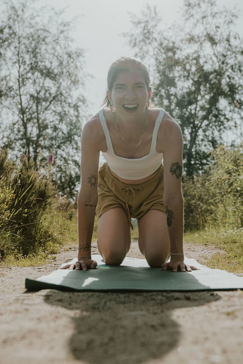 A woman practicing yoga outdoors on a sunny day, kneeling on a yoga mat, surrounded by trees and natural scenery.