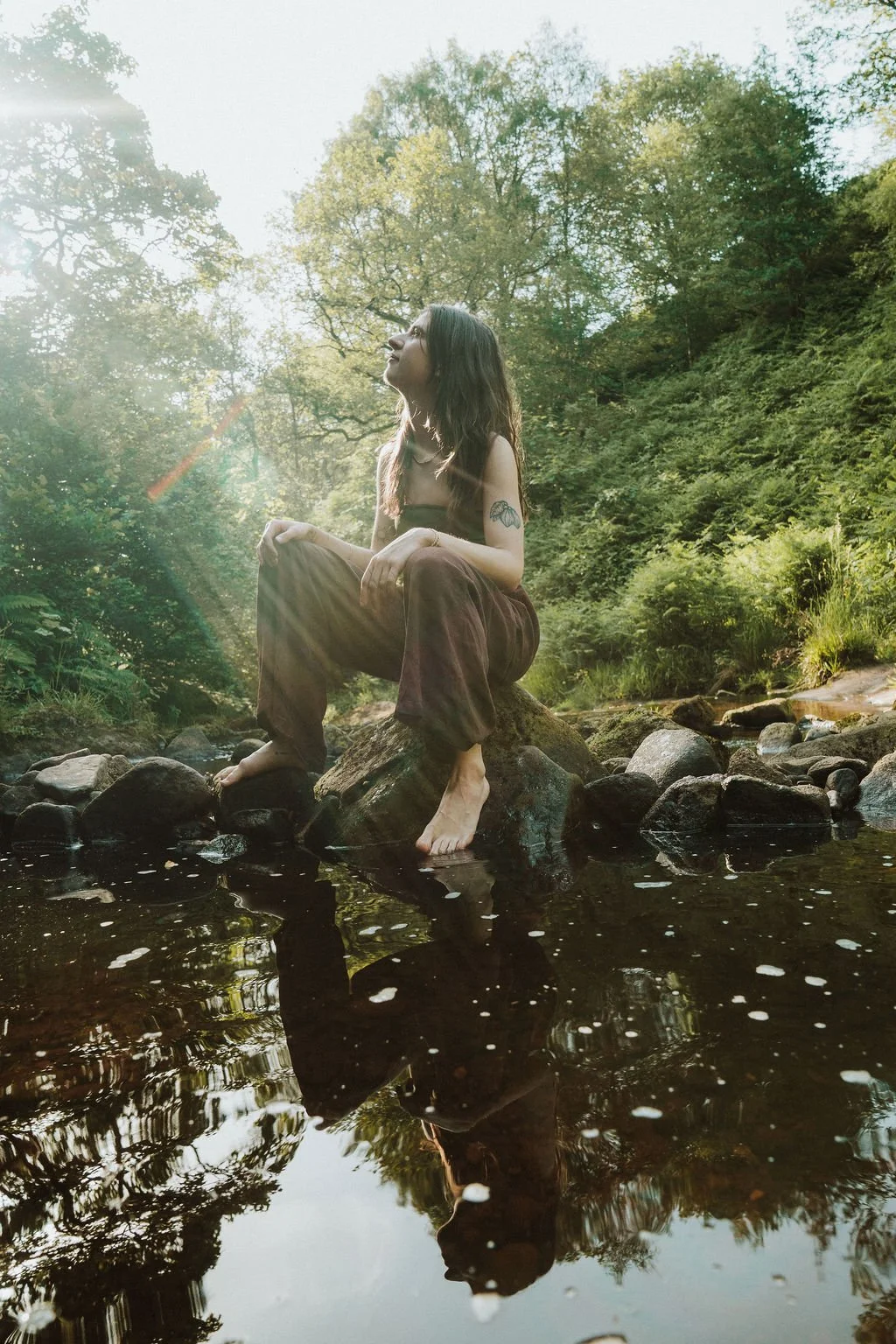 A woman sitting on a rock in a creek in a forest with sunlight filtering through trees.