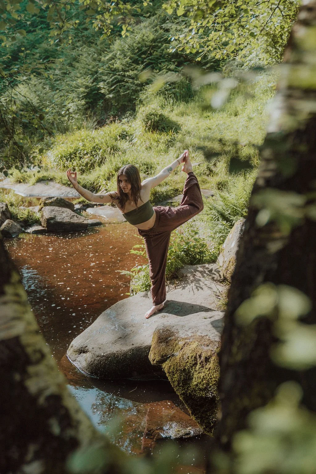 A woman practicing yoga outdoors on a rock by a small stream surrounded by lush greenery.