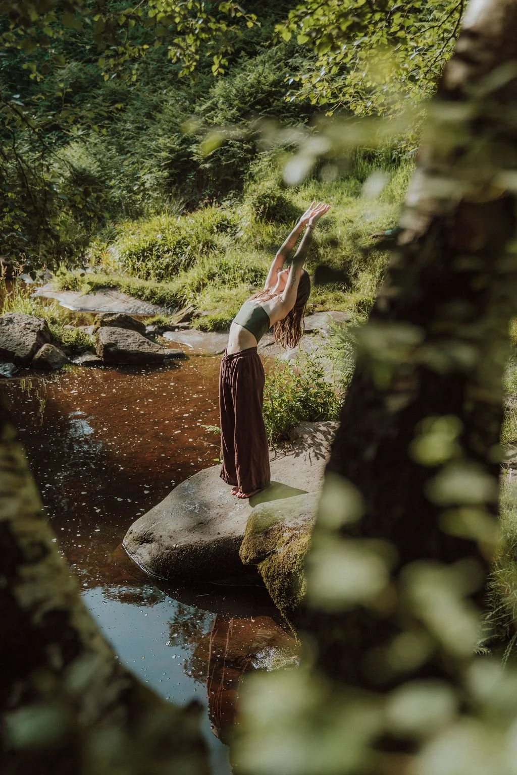 A woman doing yoga on a rock in a creek surrounded by lush green trees and plants.