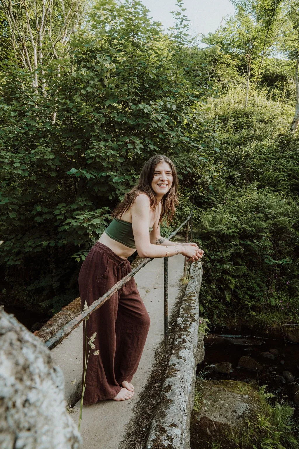 A young woman with long brown hair, wearing a green crop top and brown pants, leans on a small bridge railing smiling in a lush forest setting.