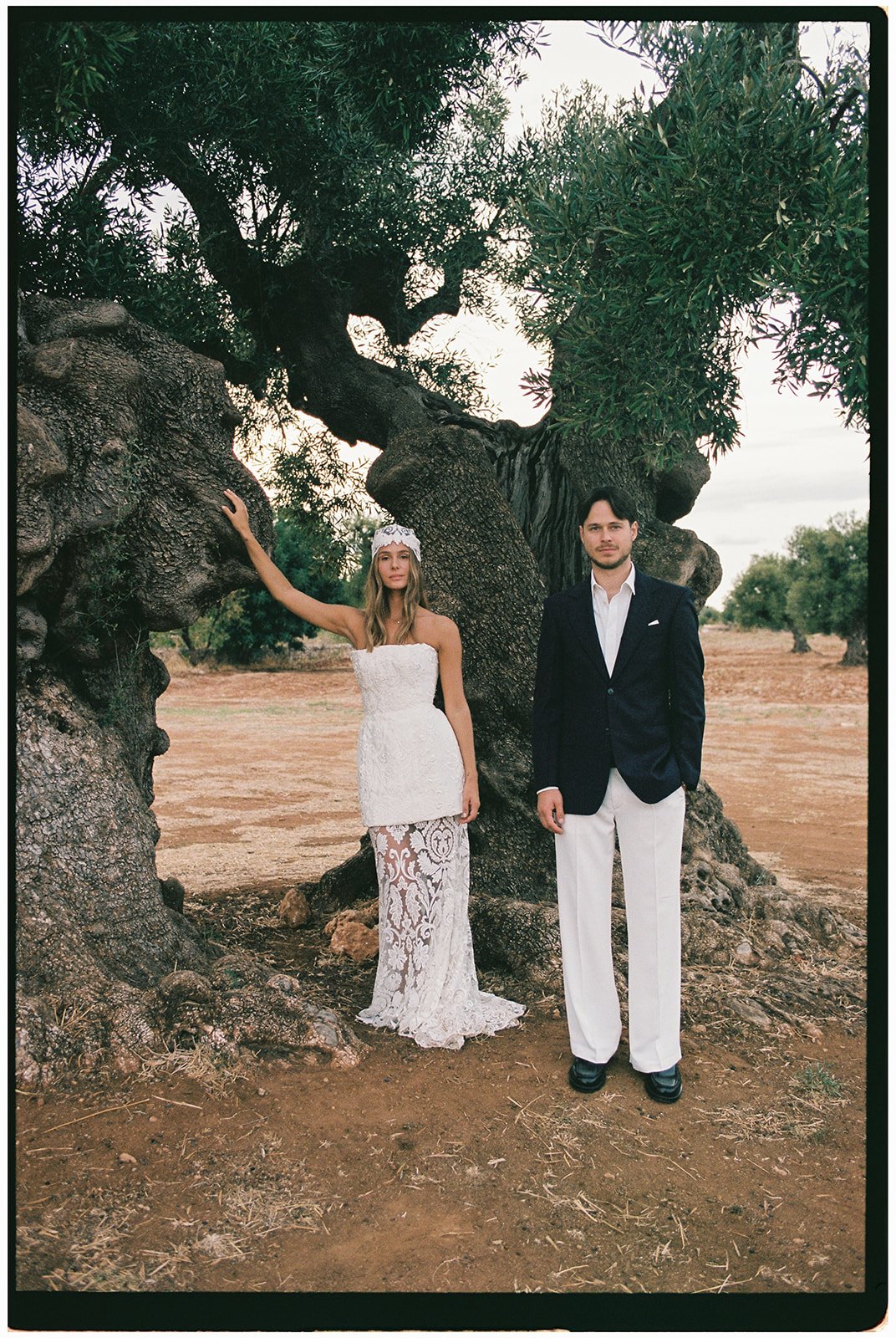 A woman in a white lace wedding dress and a man in a dark blazer and white pants stand in front of a large, twisted tree in an open, rural landscape.