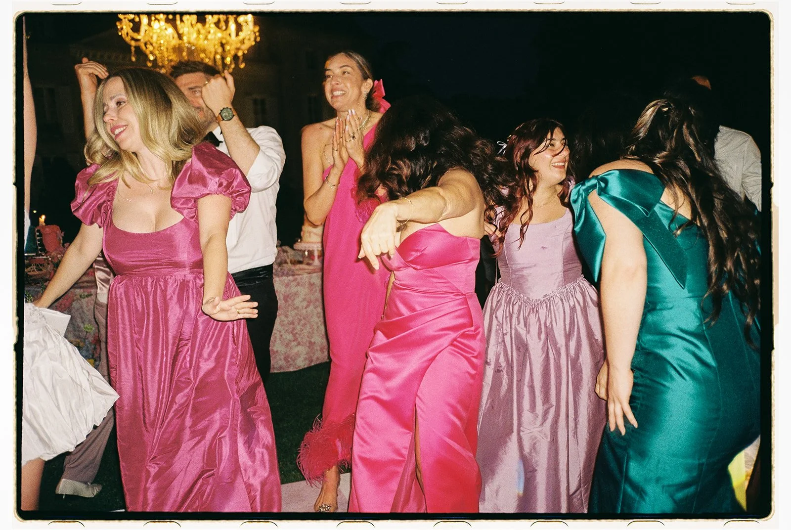 A group of women dancing and celebrating at a formal event, wearing colorful dresses, with a chandelier hanging above.