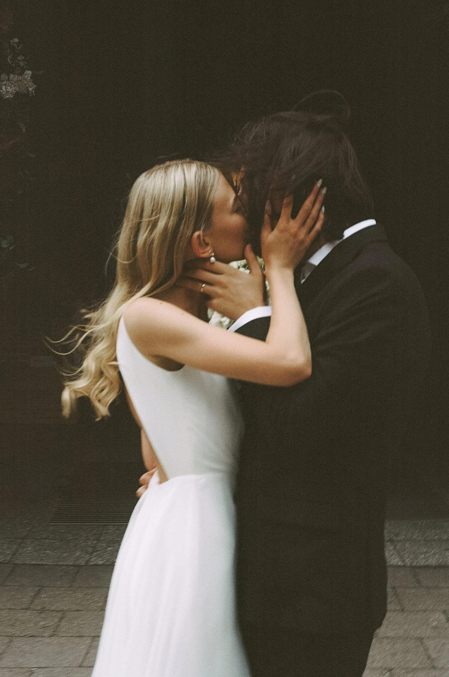 A couple engaged in a romantic kiss, with the woman wearing a white dress and the man in a black suit, outdoors at night.