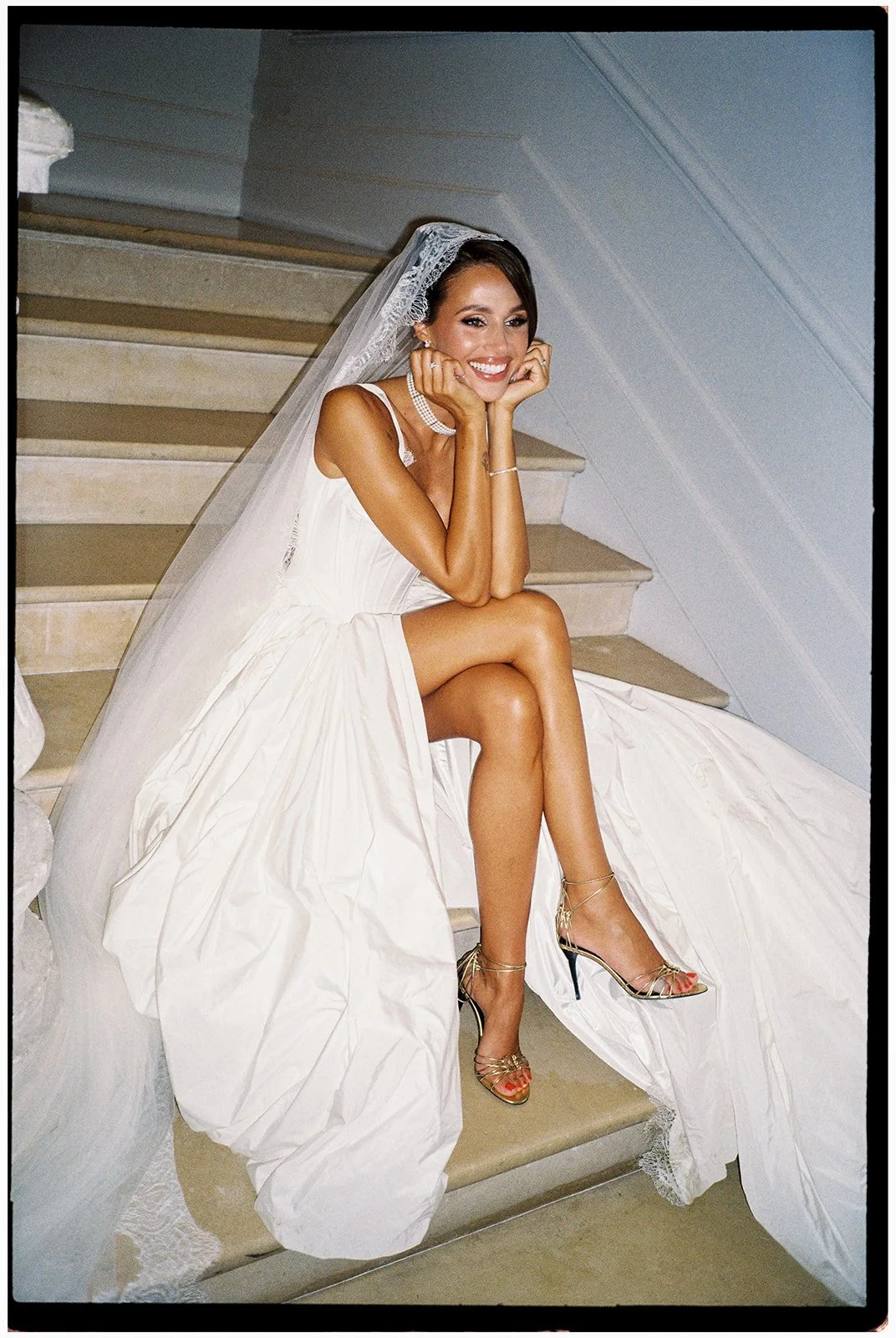 A smiling bride sitting on stairs in a white wedding dress with a veil, wearing high heels, and jewelry.