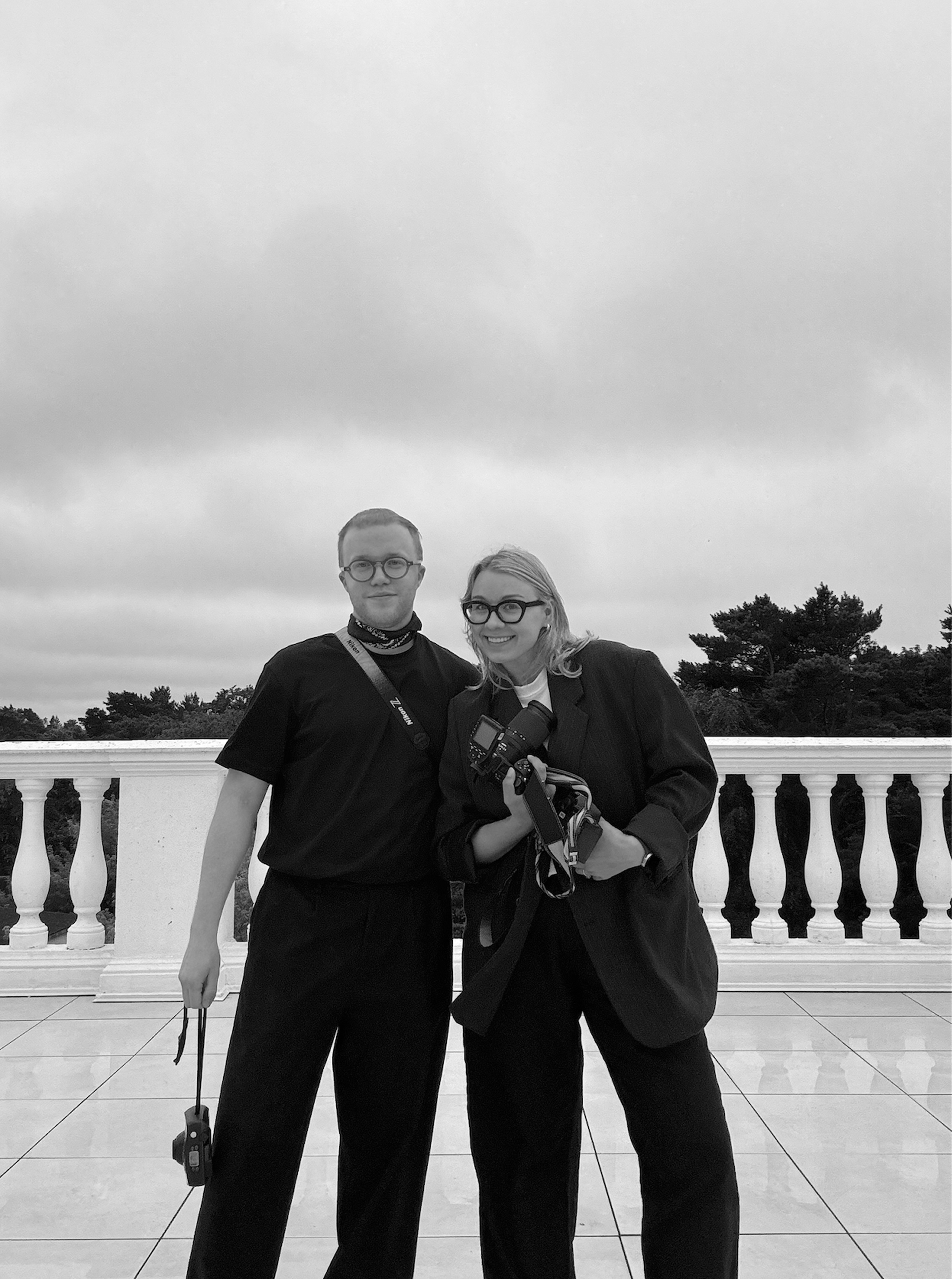 Two women standing together outdoors on a terrace with a white balustrade, holding camera equipment, both smiling and dressed in dark clothing, overcast sky overhead.