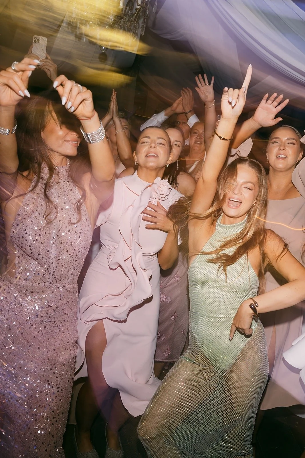Group of women dancing and celebrating at a party or celebration, wearing fancy dresses, with joyful expressions and some with raised hands.