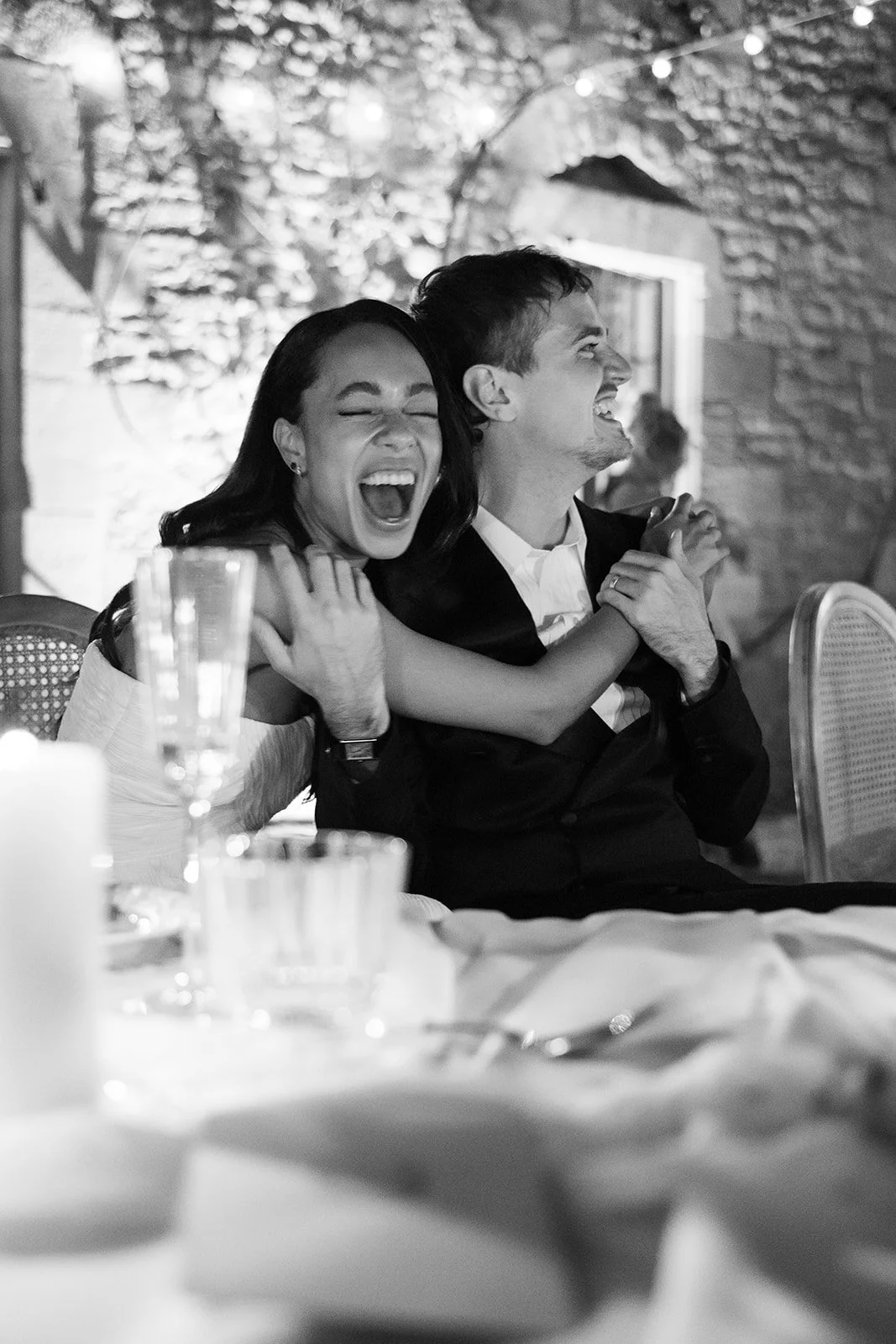 A couple enjoying a joyful moment at a celebration, sitting at a table with glasses and a candle, with a brick wall and some string lights in the background, captured in black and white.