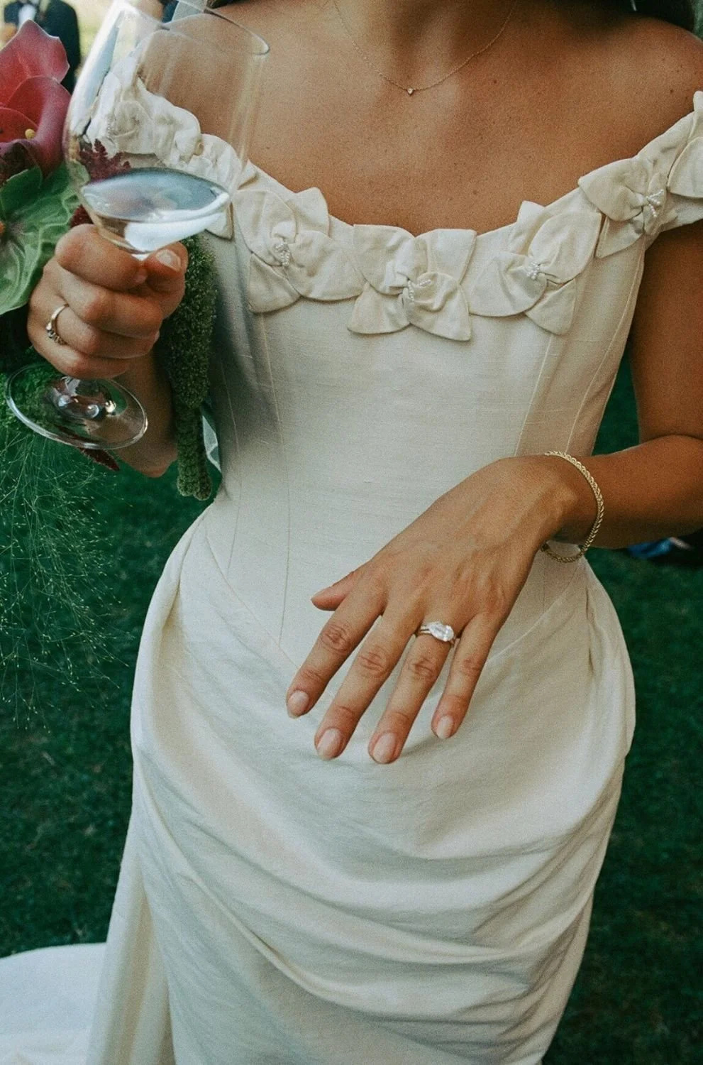 A person in a cream-colored dress holds a glass of champagne in one hand and shows off a wedding ring on her finger. She is wearing a delicate necklace and a bracelet.