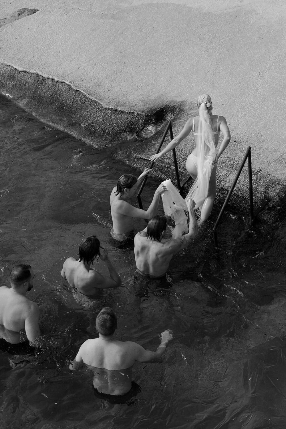 A woman in a wedding dress getting into a body of water from a concrete dock, with several men helping her, while others stand in the water watching.