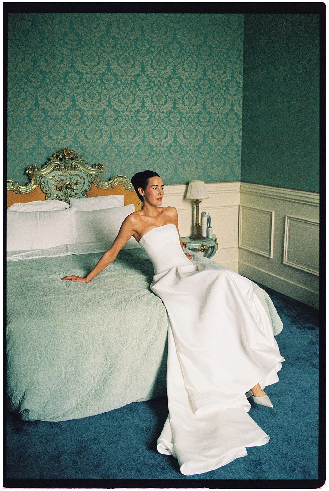 A woman in a strapless white wedding dress sitting on a bed in a Victorian-style room with blue and gold decor.