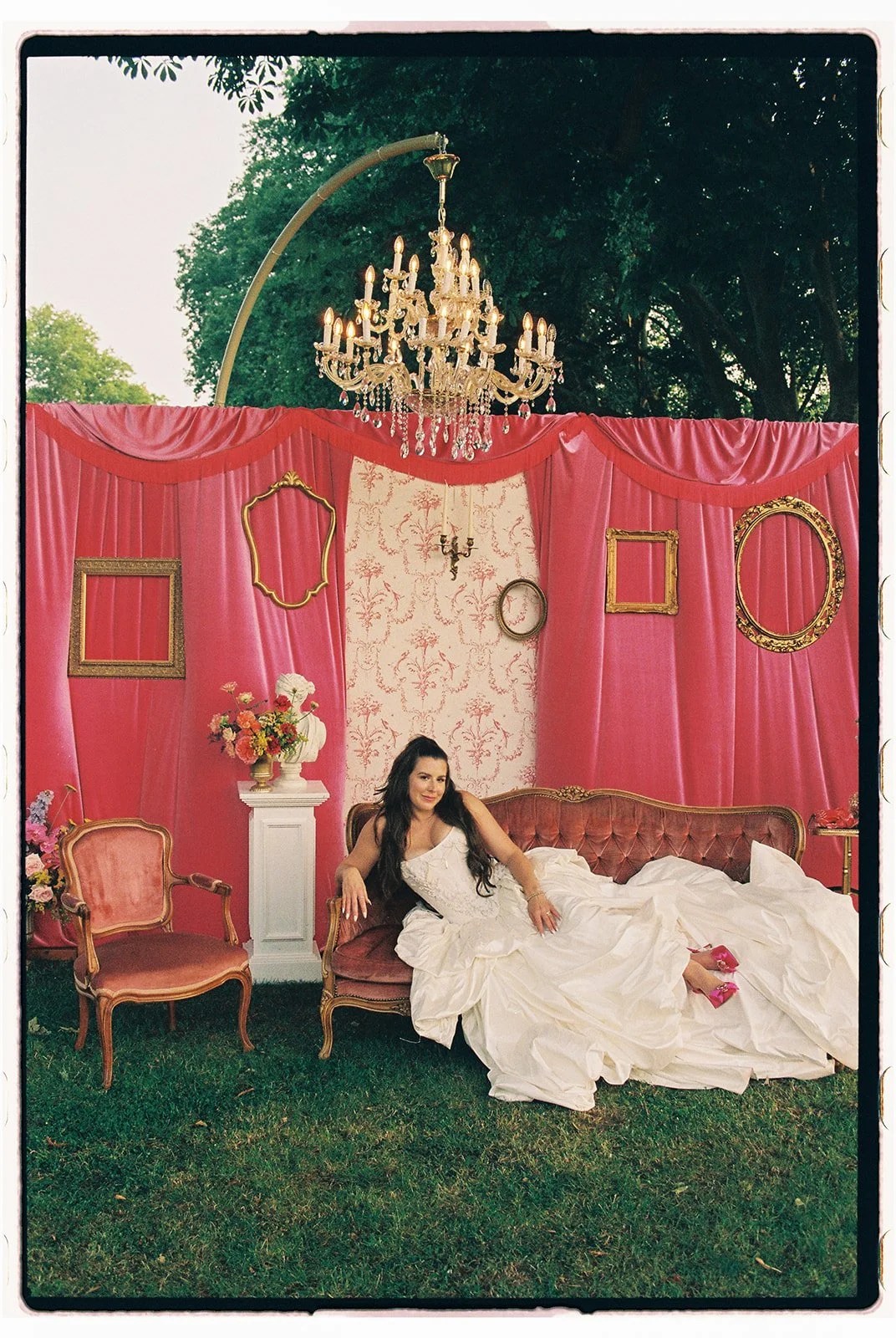 A woman in a white dress sitting on a vintage pink velvet sofa outdoors with pink curtains, gold frames, chandelier, flowers, and greenery in the background.