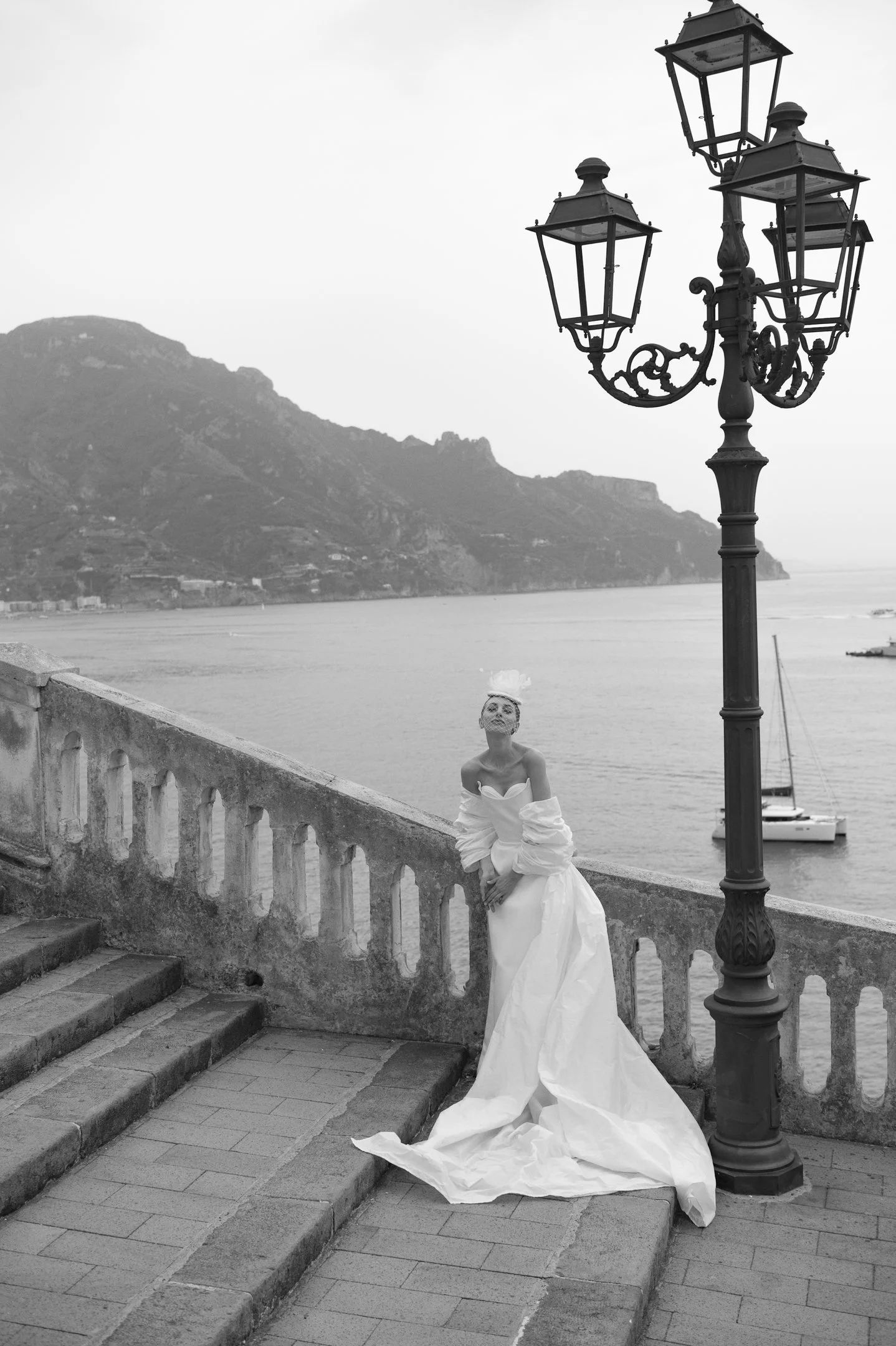A woman in a white wedding dress standing by a stone railing near water, with mountainous landscape in the background and a vintage street lamp nearby.