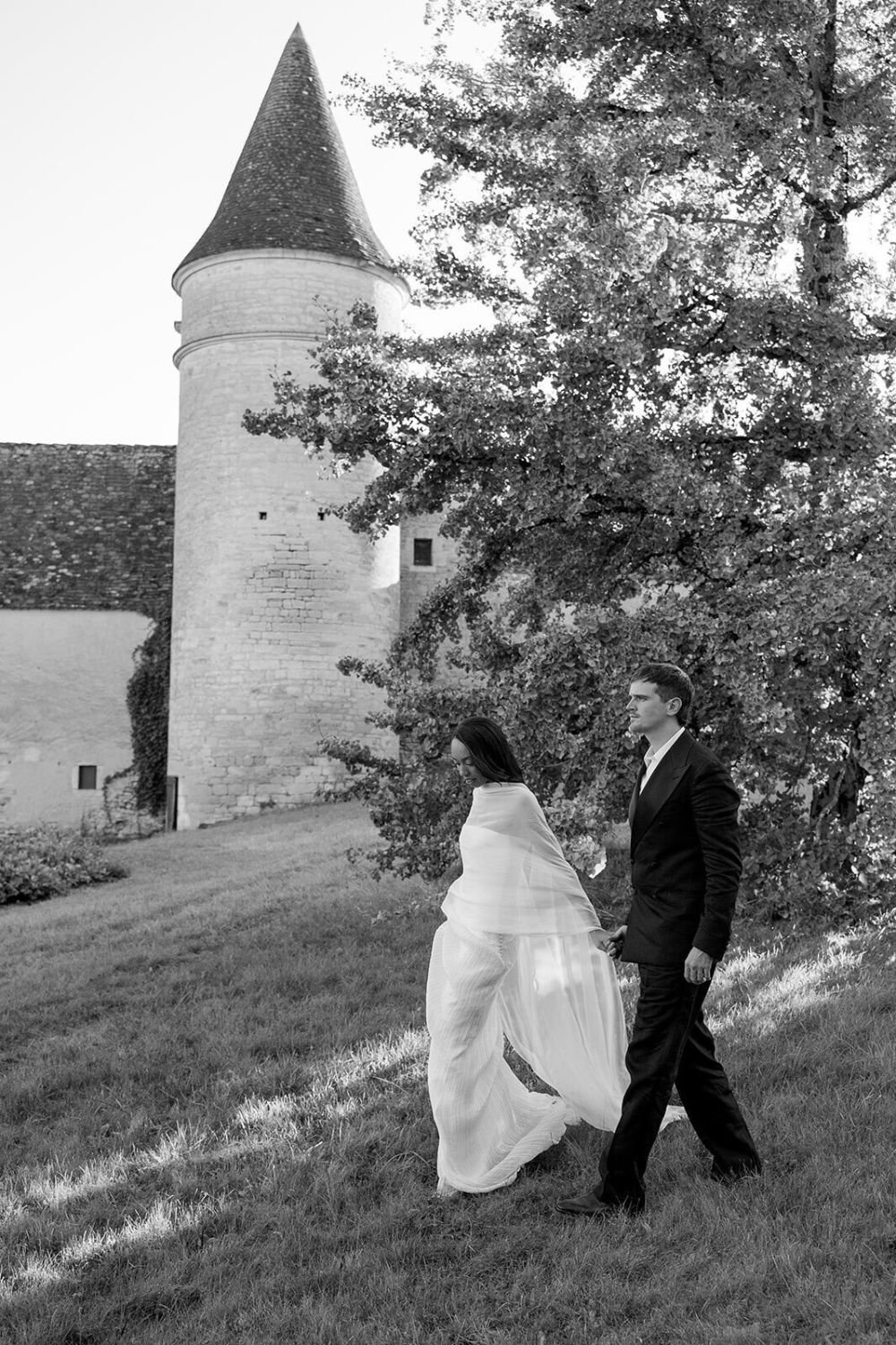 A black and white photo of a couple, a woman in a wedding dress and a man in a suit, walking together on grass near a large tree and a castle tower.