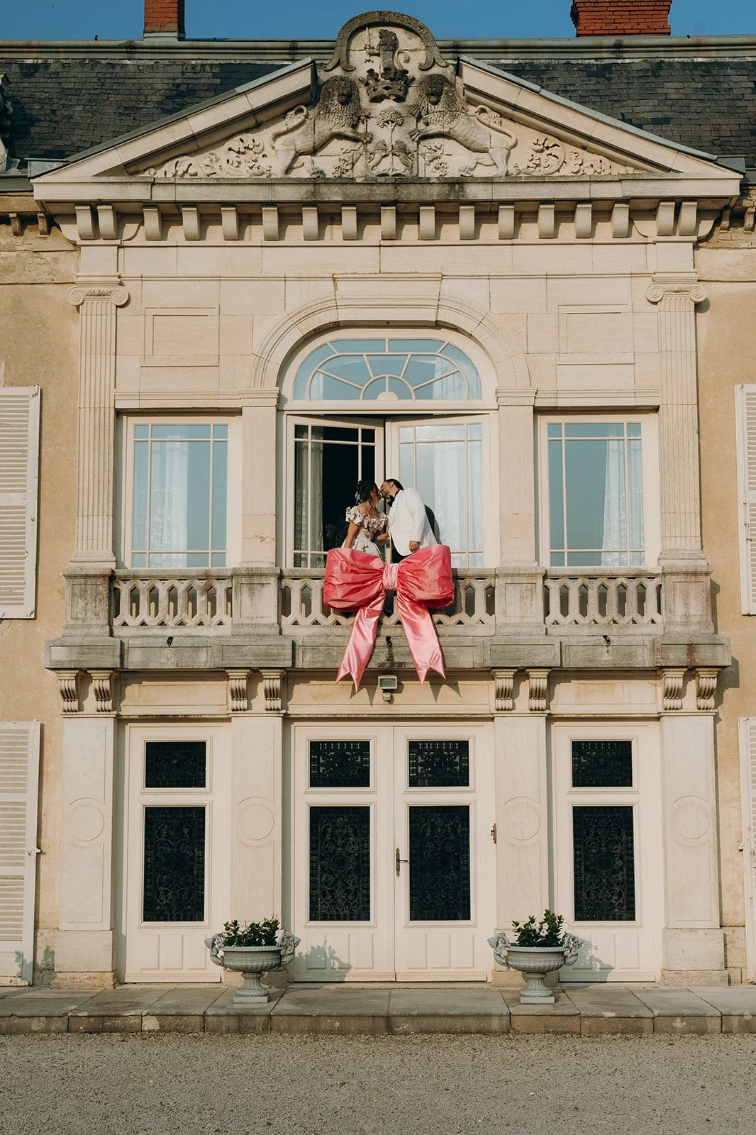 A bride and groom at a wedding, standing on a balcony of a large, historic building with a decorative crest and lion sculptures above, a large pink bow hanging from the balcony railing.