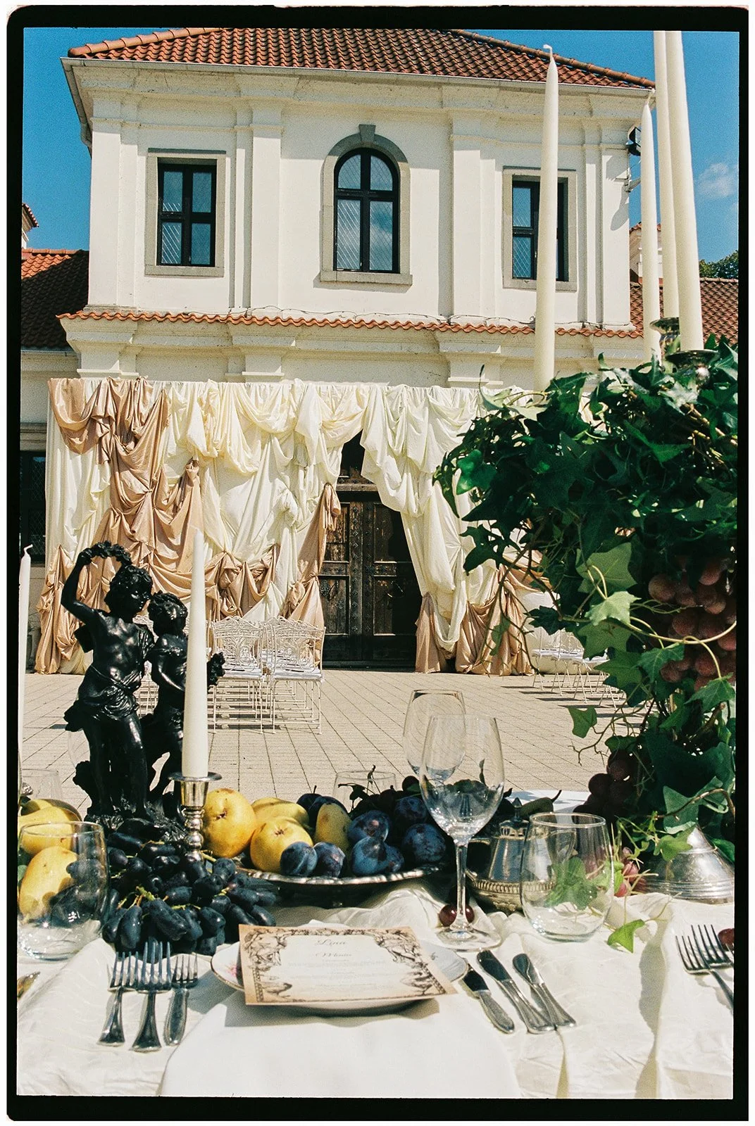 Outdoor table setting with grapes, apples, wine glasses, and a candle, overlooking a building with draped curtains and a statue.