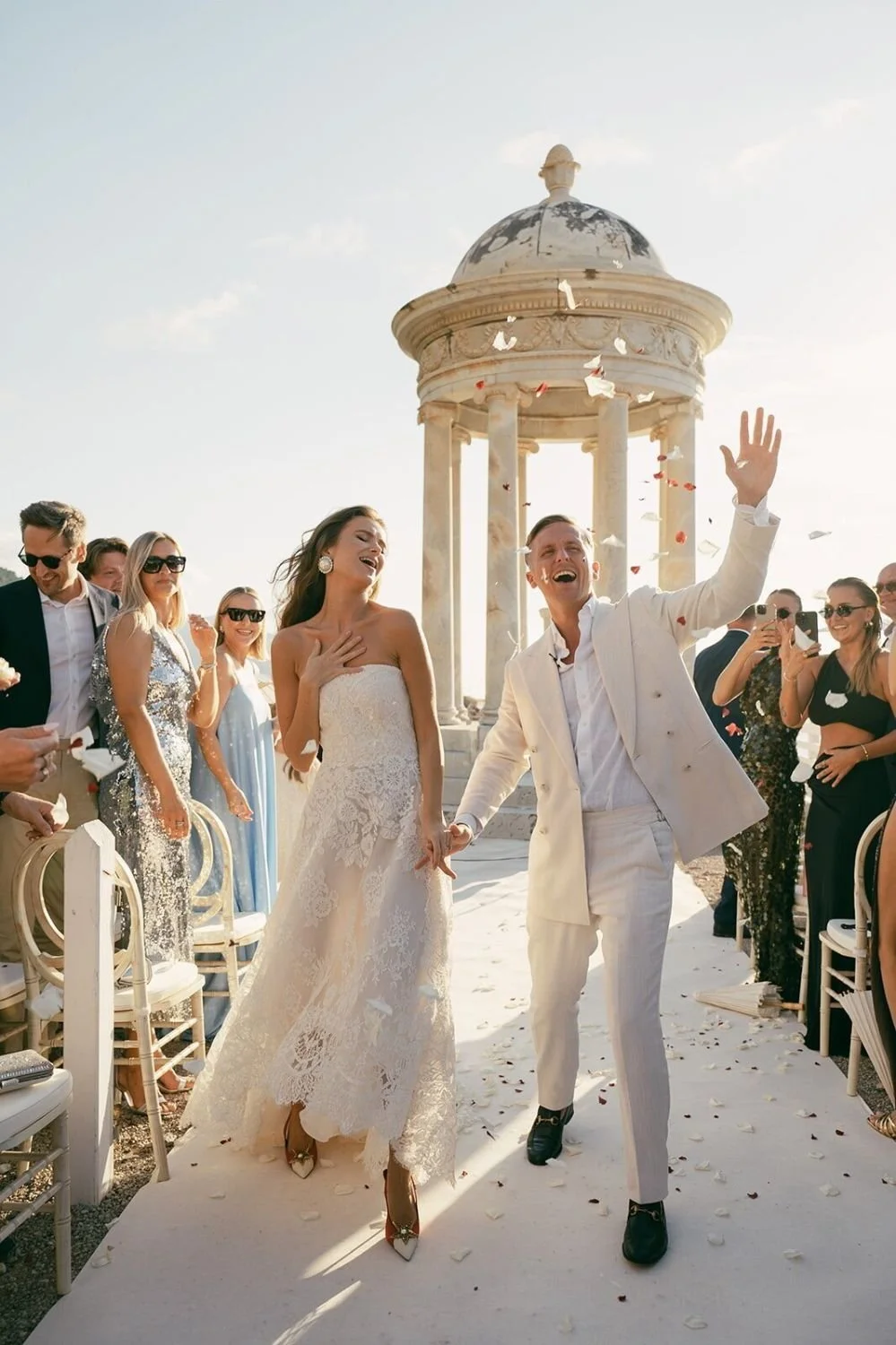 A wedding celebration outdoors with the bride and groom holding hands and smiling, surrounded by guests. The bride is in a lace wedding dress, and the groom is in a light-colored suit, both joyful. There is a stone pavilion in the background and flow