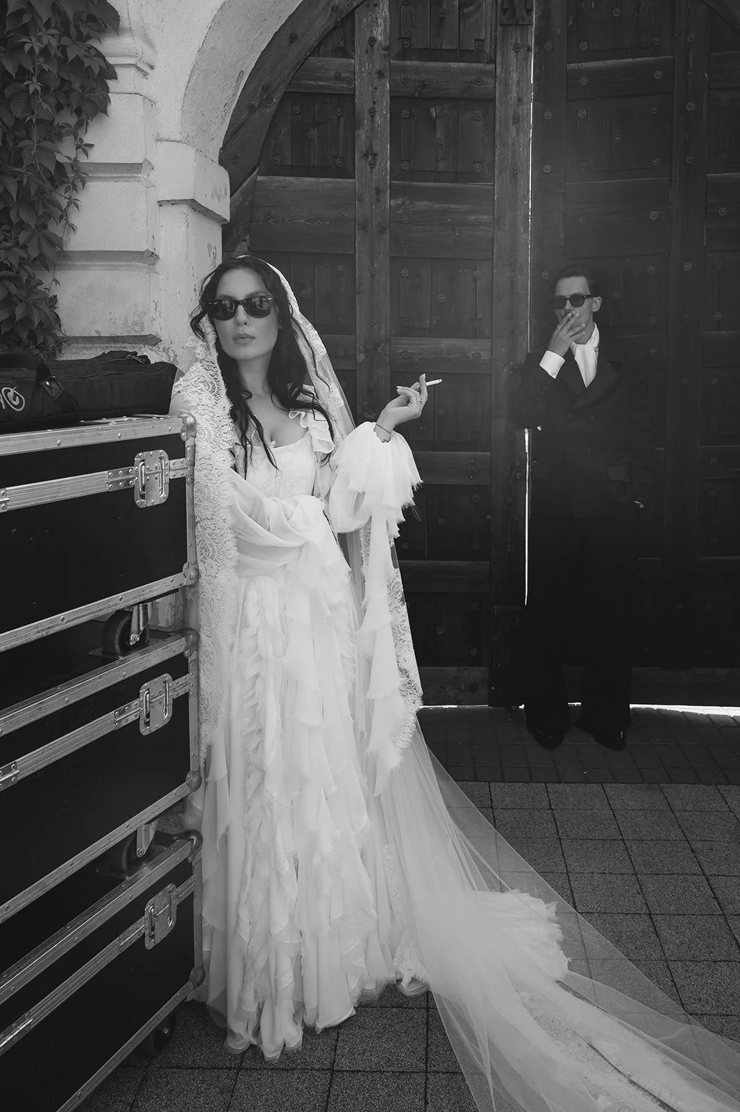 A woman in a wedding dress and sunglasses standing next to a man in a dark suit and sunglasses, smoking a cigarette, in front of large wooden doors.