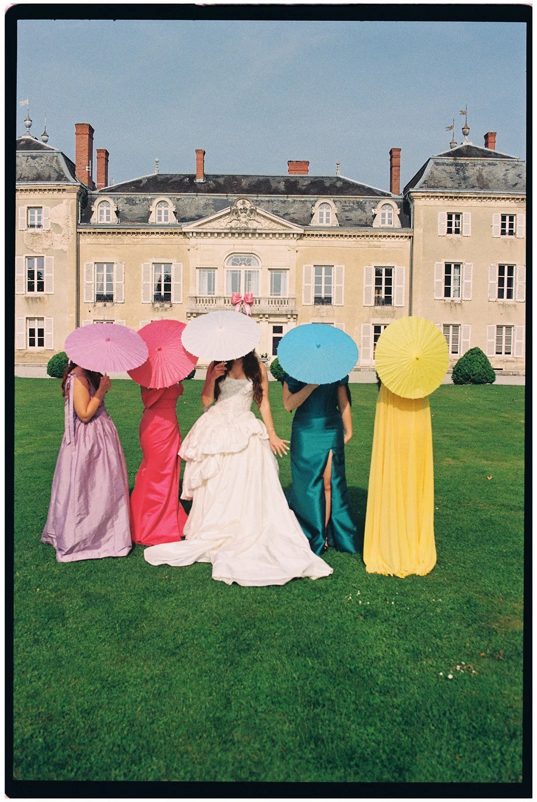 Five women in colorful evening gowns holding umbrellas and standing on a lawn in front of a historic mansion.