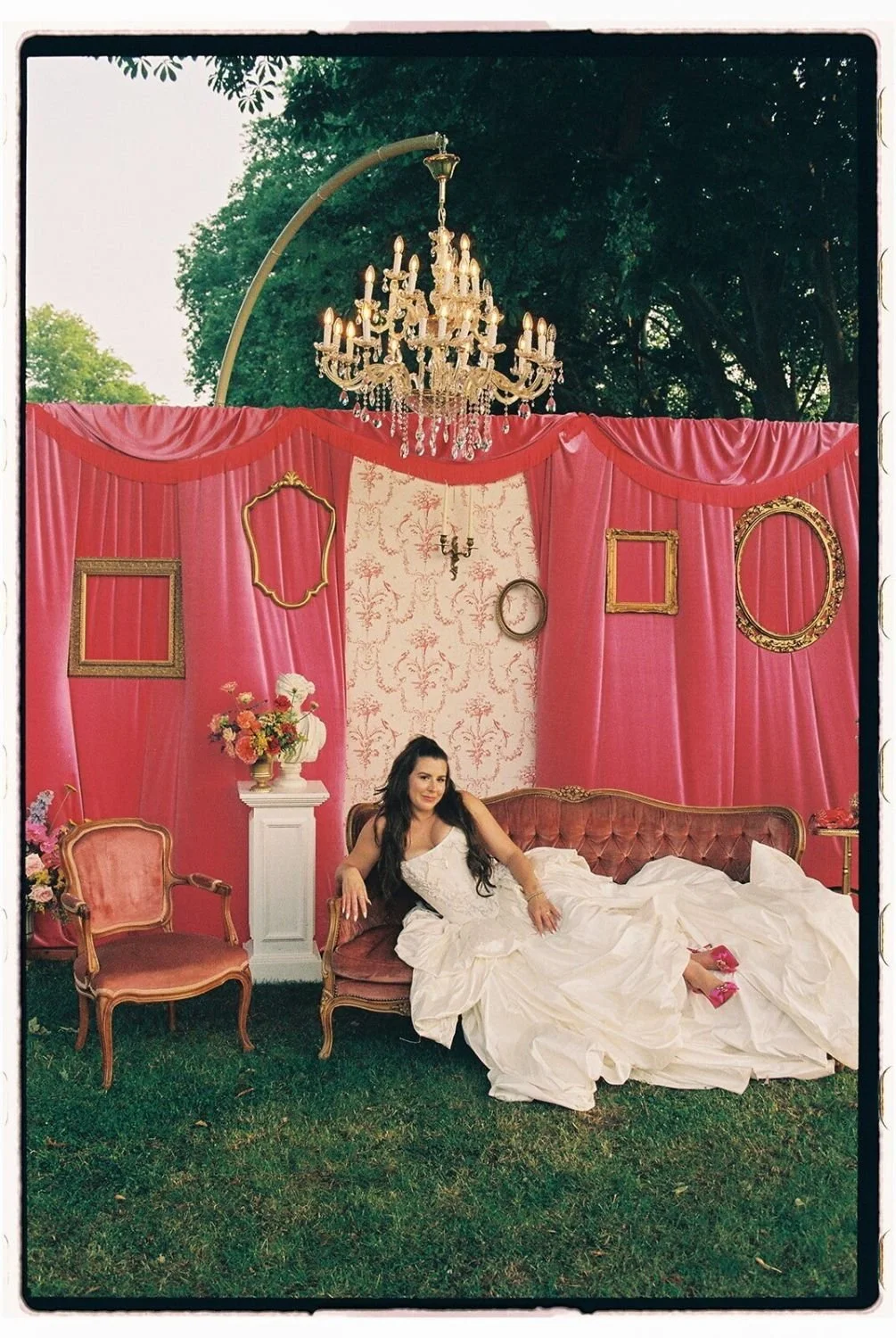 Woman in white dress lying on a vintage pink sofa in a decorated outdoor setting with pink drapes, gold frames on the wall, a chandelier above, and floral arrangements.