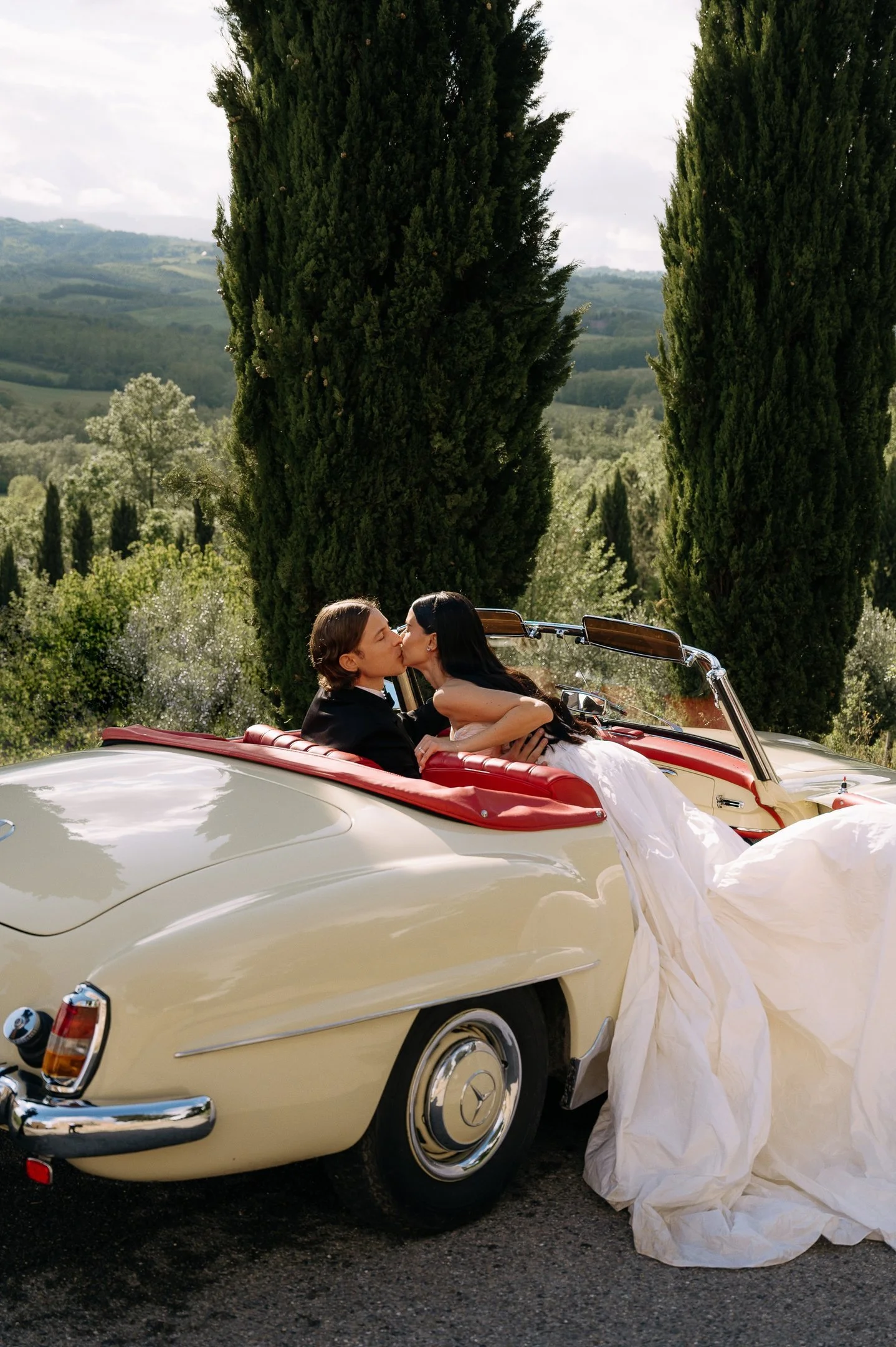 A newlywed couple sharing a kiss in a beige vintage Mercedes-Benz convertible with a white wedding dress draped over the side, set against a scenic landscape of tall cypress trees and rolling hills.