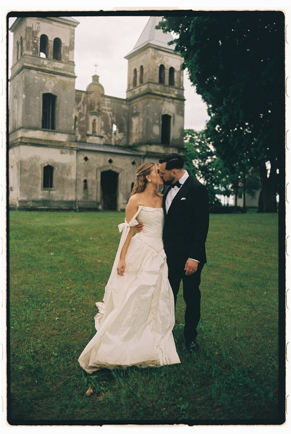 A bride and groom are sharing a kiss in front of a historic church with twin towers. The bride is wearing a white wedding gown and the groom is in a black tuxedo.