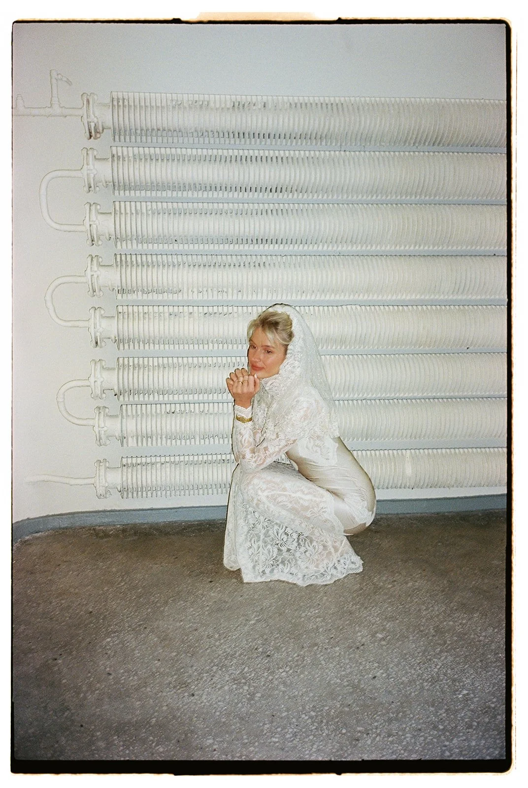 A woman dressed in white lace clothing crouching in front of a wall with a painted mural of large, white utility pipes.