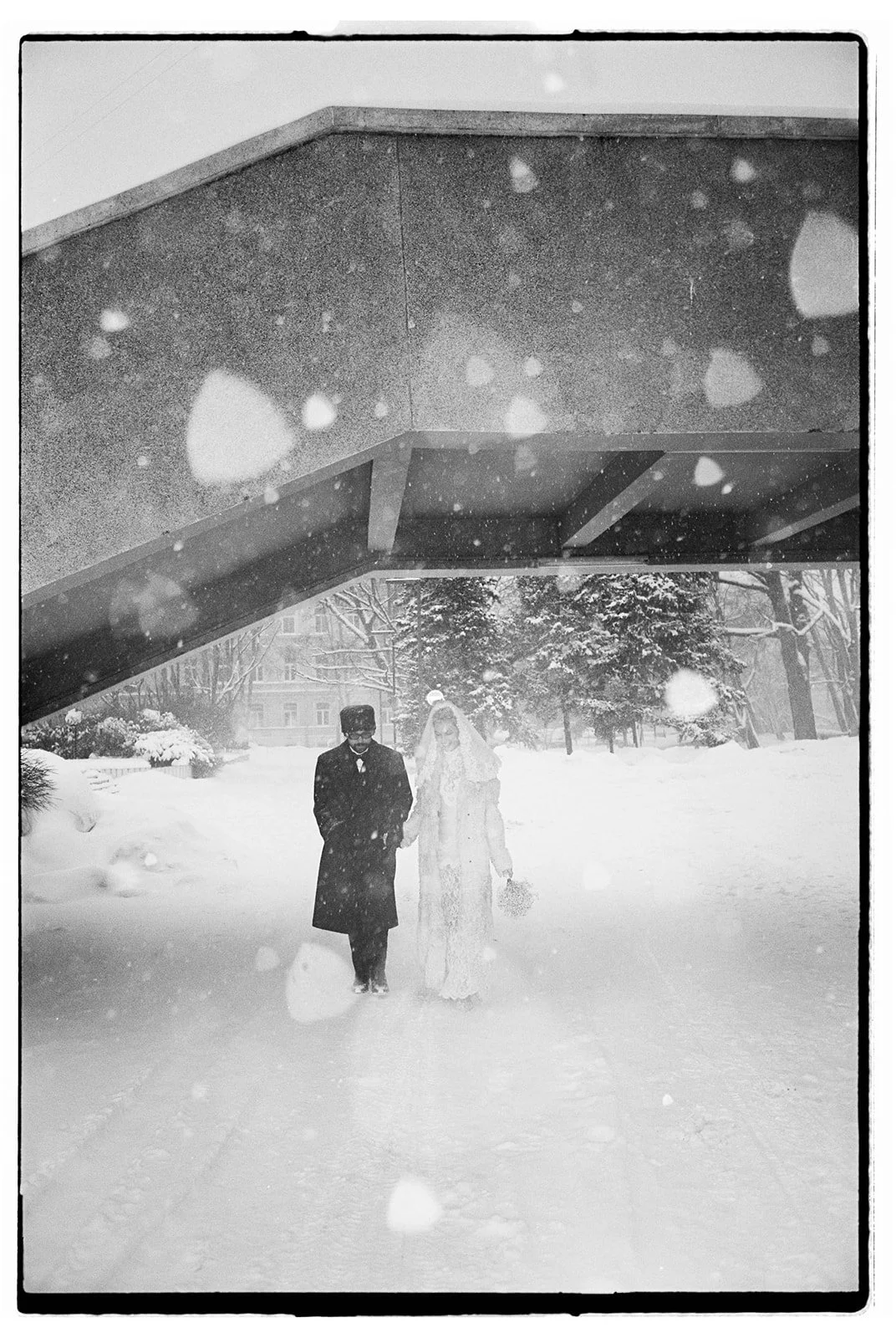 Black and white photo of a bride and groom walking in the snow outdoors, under a large concrete bridge, with snow-covered trees and buildings in the background.