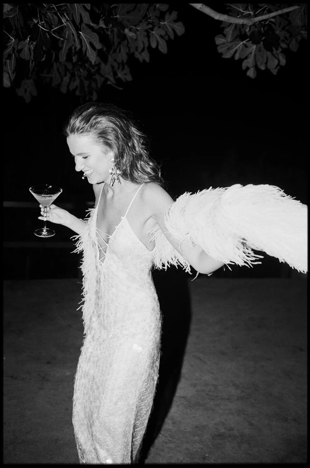 A woman in a white, feathered dress holding a martini glass, smiling, with long earrings, and a feathered accessory on her arm, standing outdoors at night.