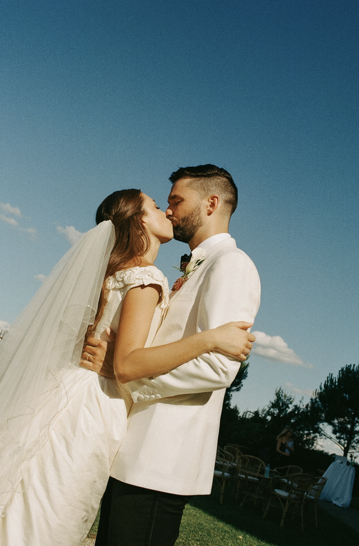 A bride and groom sharing a kiss outdoors on their wedding day under a clear blue sky.