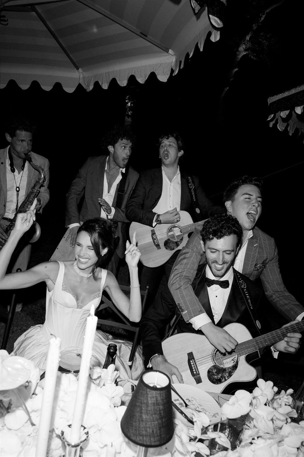 Group of people at a celebration, some playing musical instruments, others singing and dancing, with candles and flowers on the table.