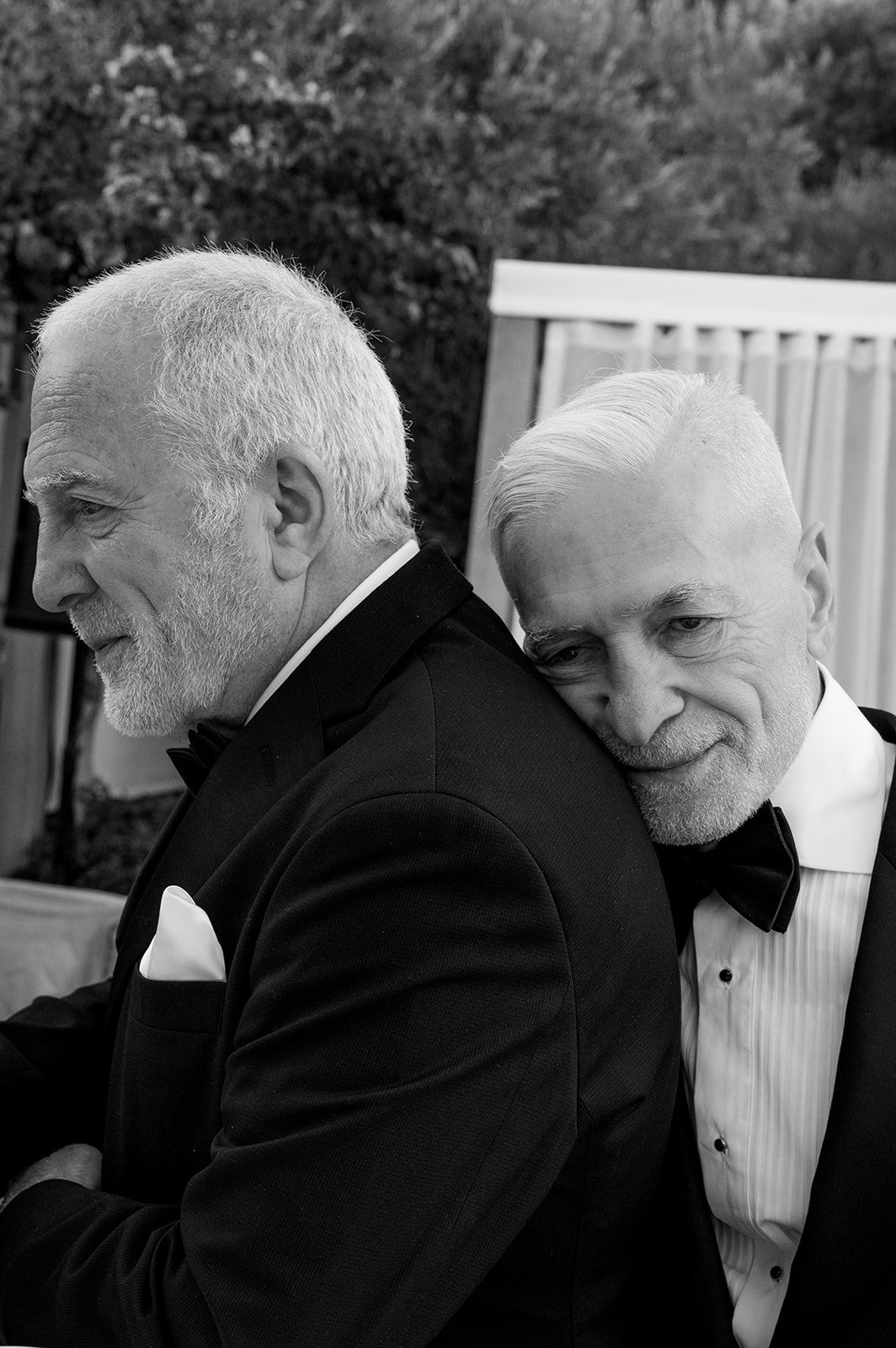 Two elderly men dressed in tuxedos, one with a beard and the other with a clean face, sharing an intimate moment at a formal event.