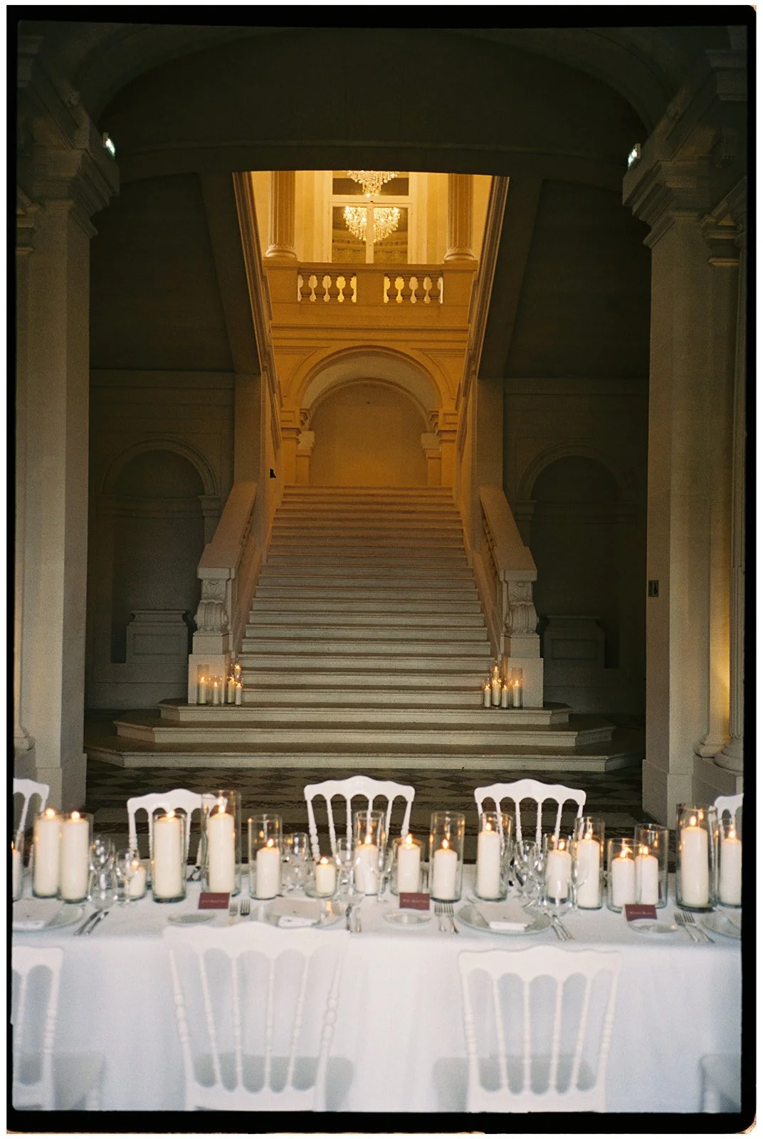 Elegant dining table set with white tablecloth, candles, and glassware in front of a grand staircase inside a luxurious building with chandeliers.