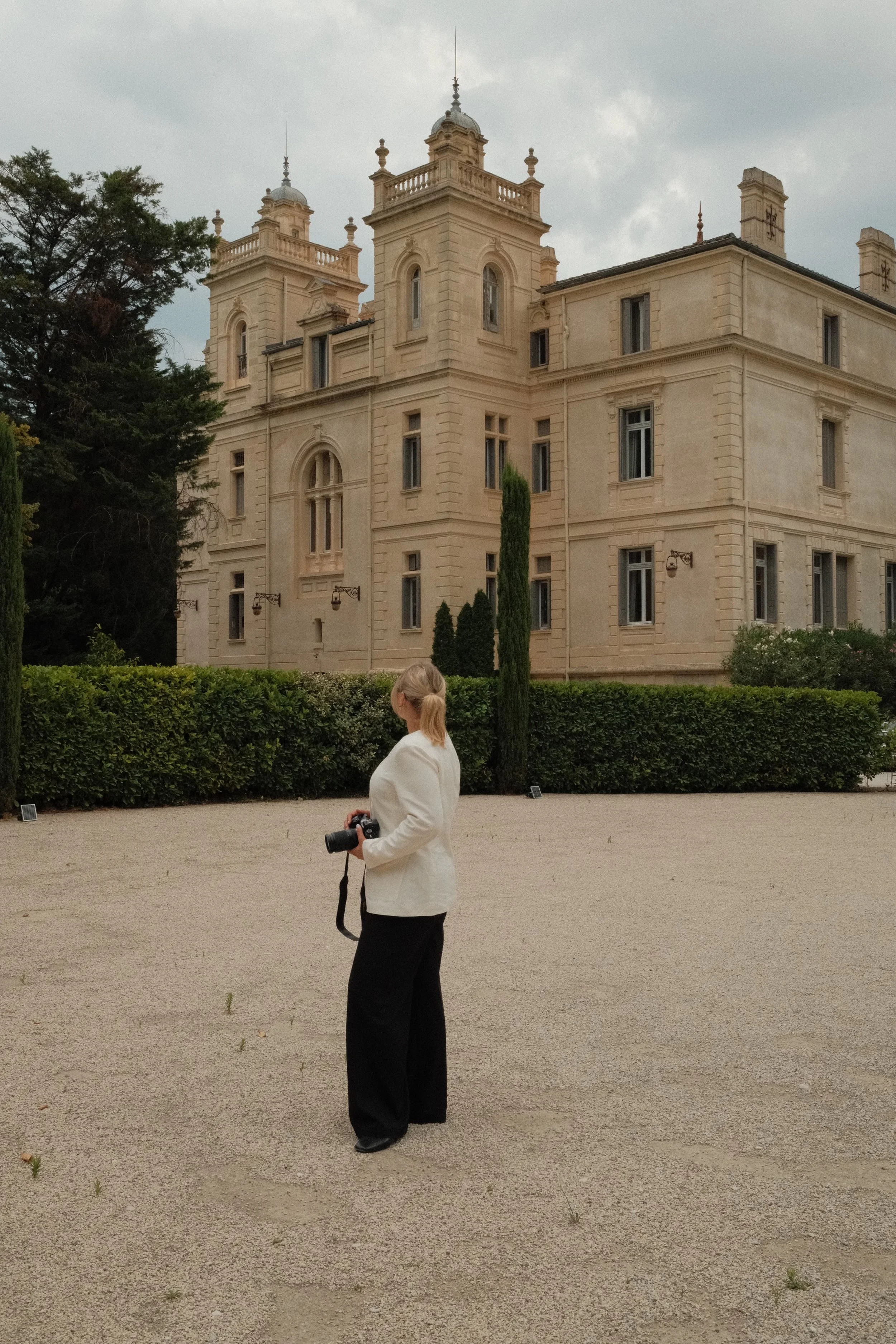 A woman with blonde hair in a ponytail, wearing a white blazer and black pants, stands outdoors holding a camera. Behind her is a large, historic, beige stone building with towers and lush green landscaping, under a cloudy sky.