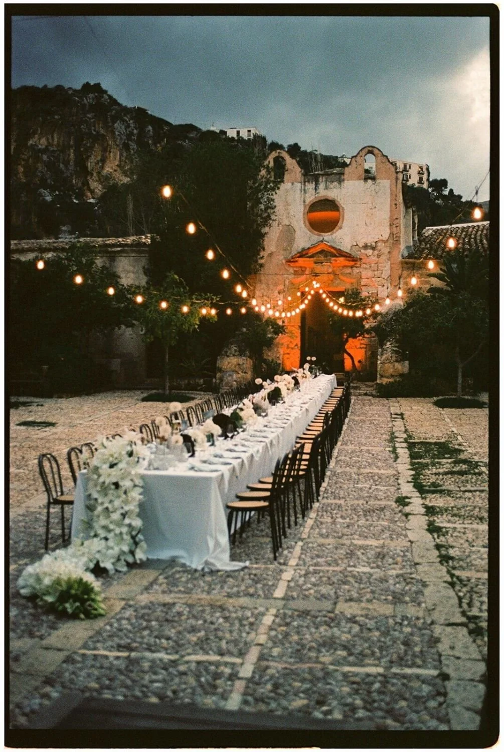 Outdoor wedding reception setup with a long white-draped table decorated with white floral arrangements, string lights overhead, and an old stone building with a circular window in the background at dusk.