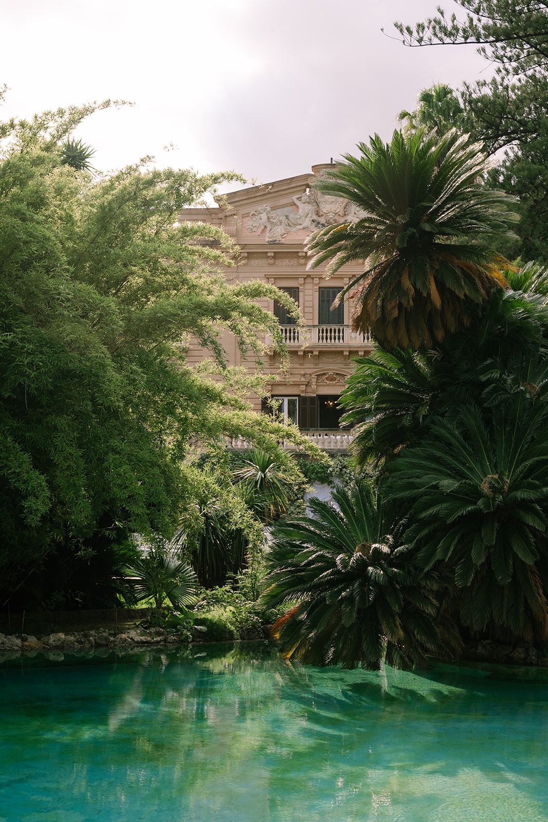 Historical building with classical architecture surrounded by lush green trees and large palm plants, with a pool of water in the foreground.