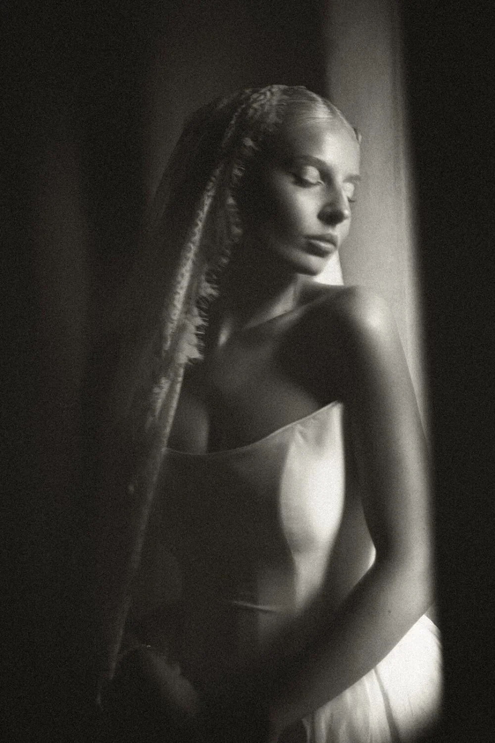 A black and white photo of a woman with styled hair and closed eyes, sitting near a window, illuminated by soft light.