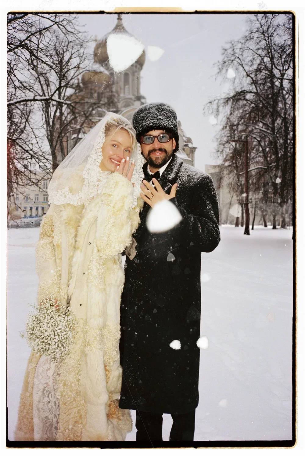 A bride and groom in winter clothing standing outdoors in the snow, with a church with onion domes in the background. The bride is wearing a cream-colored fur coat and veil, holding a bouquet of white flowers, while the groom is wearing a black coat 