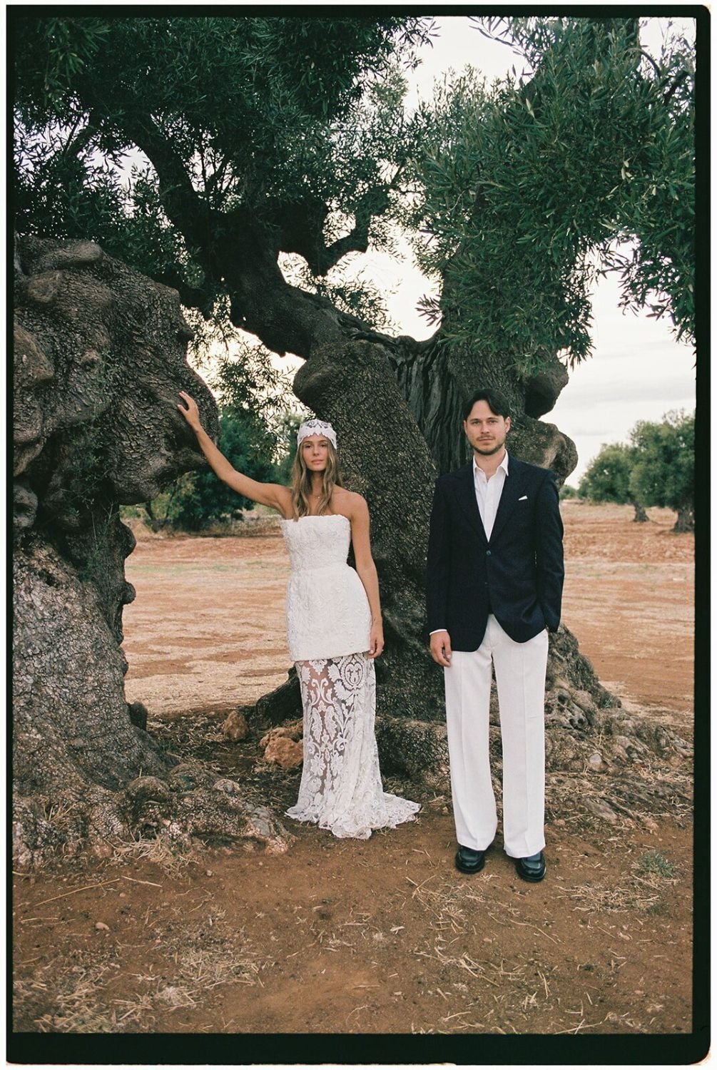 A woman in a white lace wedding dress and a man in a suit standing outdoors in front of a large, twisted tree with green leaves, on a dirt ground with an overcast sky in the background.