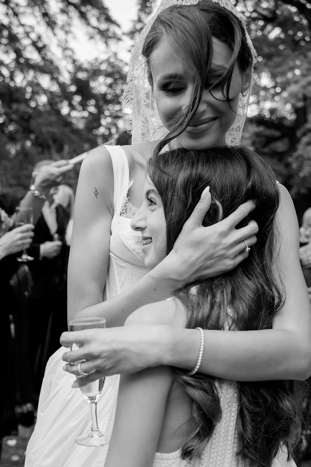 Two women in wedding dresses hugging at an outdoor wedding celebration, one woman with dark hair smiling, the other with light hair and veil, background with trees and guests