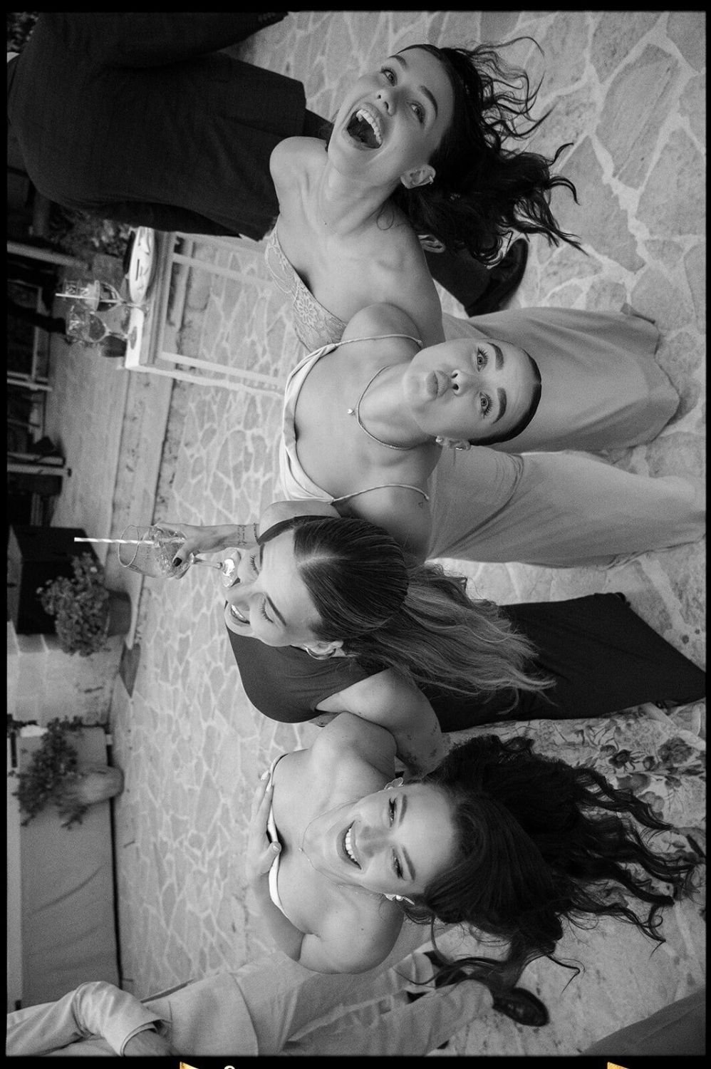 Group of five women at a celebration, making playful faces and sticking out their tongues, in an indoor setting with a stone floor and tables in the background.