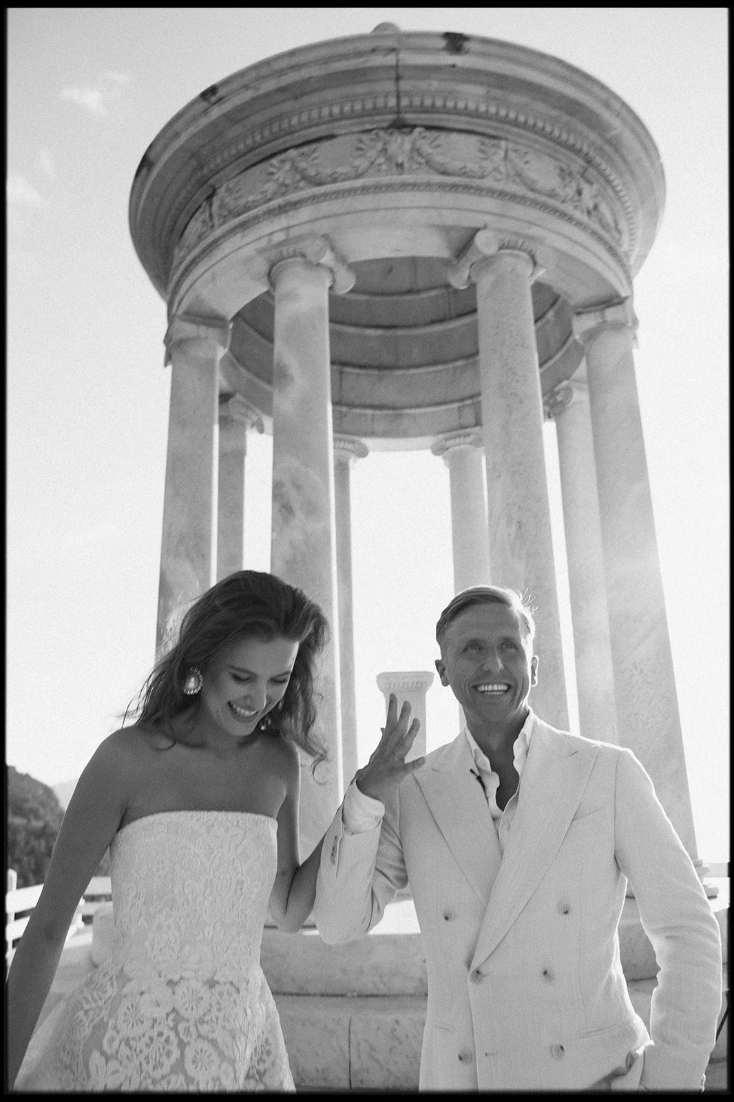 Black and white photo of a smiling couple in wedding attire standing in front of a classical Greek-style pavilion with columns.