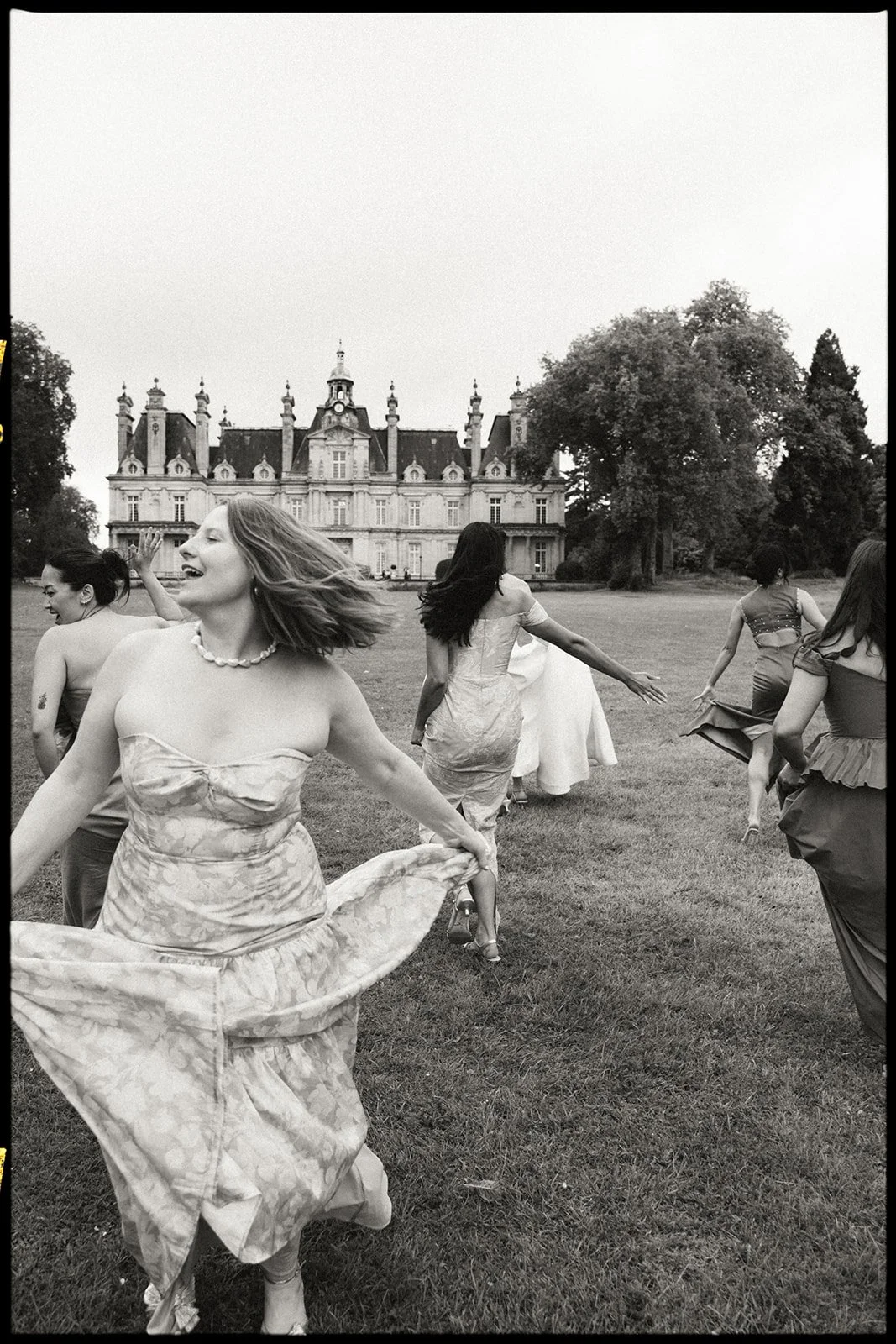 Women in elegant dresses dancing and playing on a lawn in front of a castle.