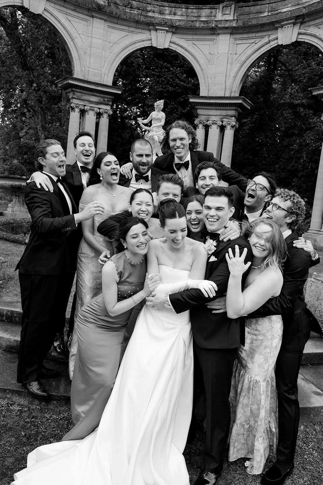 Group of people dressed in formal suits and dresses, celebrating and smiling outdoors in front of architectural arches and statues.
