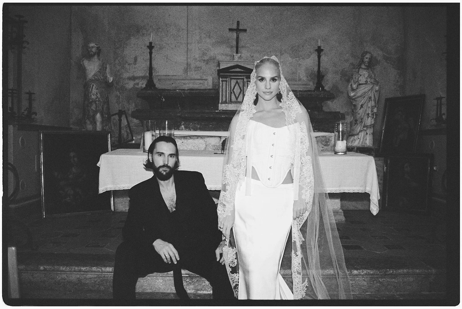 Black and white photo of a woman in a wedding dress standing next to a seated man in a church or chapel setting, with religious statues and altar in the background.
