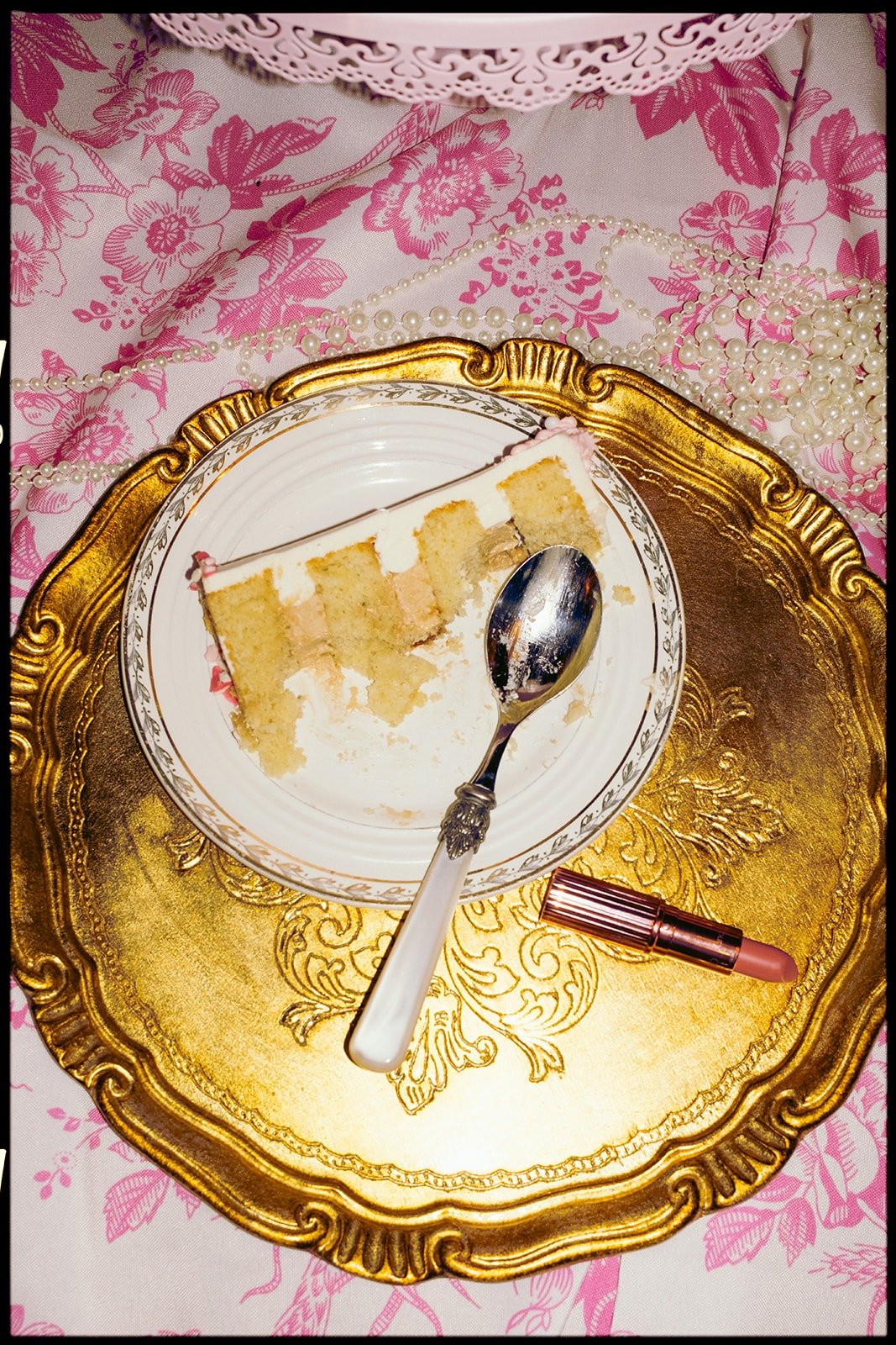 A partially eaten slice of cake with pink icing on a decorative white plate with a gold rim, placed on an ornate gold tray. There is a silver spoon with a white handle, a lipstick, a string of pearls, and a pink floral patterned cloth in the backgrou
