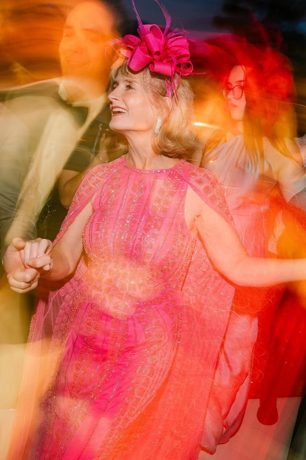 A smiling woman in a pink, embellished dress with a large matching pink bow headpiece, dancing at a lively celebration or party.