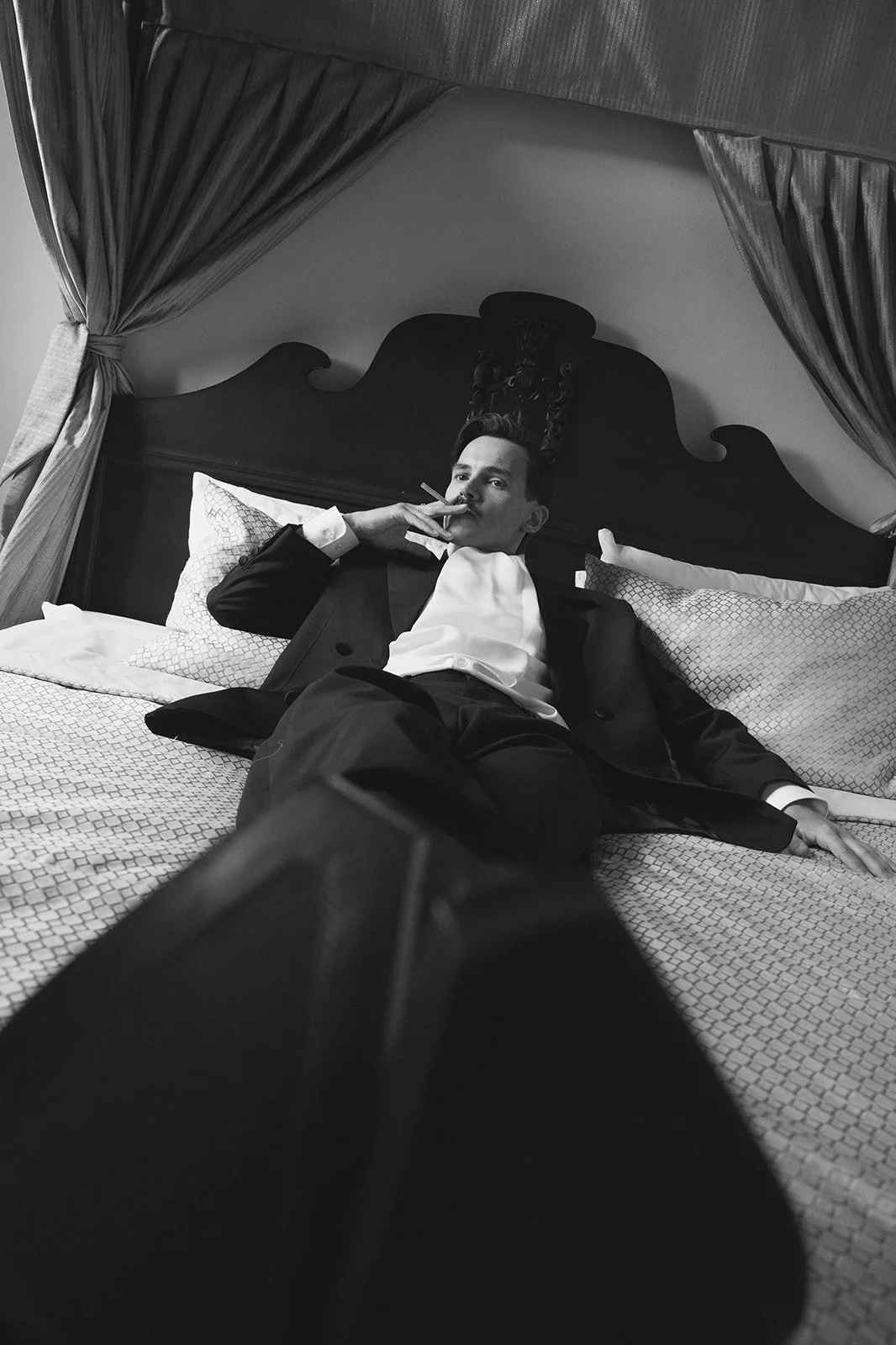 A man dressed in a tuxedo reclining on a bed, smoking a cigarette, with a relaxed pose in a hotel room with curtains and pillows.