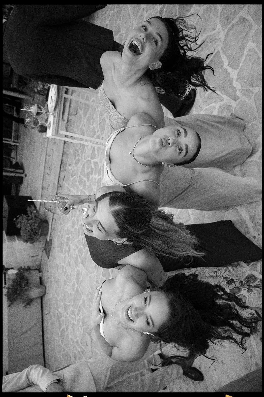 Group of five women gathered around a table, making playful faces and sticking out their tongues, at a social event or party.